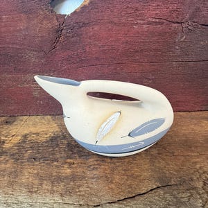 May include: A cream-colored ceramic pitcher with a smooth, rounded body and an integrated handle. The exterior features painted gray feather motifs and a horizontal gray band near the base. The interior of the spout is a soft gray color.