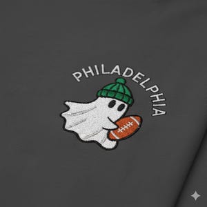 Puede incluir: Una tela gris oscuro con un diseño bordado de un fantasma blanco que lleva un gorro verde y sostiene un balón de fútbol americano. La palabra PHILADELPHIA está bordada en letras mayúsculas blancas en un arco sobre la figura.