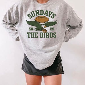 May include: A heather grey crewneck sweatshirt featuring a graphic print with the text SUNDAYS ARE FOR THE BIRDS in dark green. The design includes a brown football above a green eagle with outstretched wings and a small brown football icon.