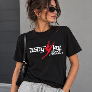 May include: A black short-sleeved t-shirt featuring a white graphic logo that reads "abby lee" with the words "DANCE COMPANY" underneath. A red silhouette of a dancer in a split leap pose is positioned between the words "abby" and "lee".