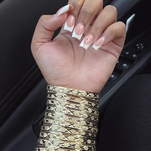 May include: A hand with French-manicured nails, featuring white tips and a nude base, adorned with a small design on one nail. The wrist is stacked with multiple gold bangles, each displaying a textured, geometric pattern. The overall aesthetic is elegant and stylish.