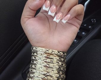 Könnte beinhalten: Eine Hand mit French-Maniküre, mit weißen Spitzen und einer nackten Basis, verziert mit einem kleinen Design auf einem Nagel. Das Handgelenk ist mit mehreren goldenen Armreifen geschmückt, die jeweils ein strukturiertes, geometrisches Muster aufweisen. Die Gesamtästhetik ist elegant und stilvoll.
