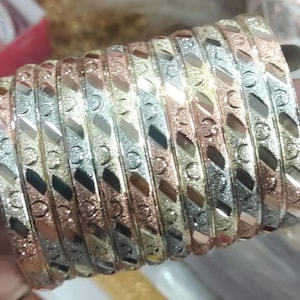 May include: A stack of tri-color bangles. The bangles are made of gold, silver, and rose gold. Each bangle has a textured surface with a repeating geometric pattern. The bangles are stacked together, showcasing the different colors and designs.