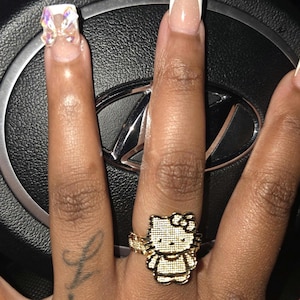 May include: A gold-colored ring featuring a Hello Kitty design, worn on a finger with a French manicure. The ring is detailed with a bow and the iconic character's face. The nails have a clear base with white tips. The ring is on a finger with a tattoo.