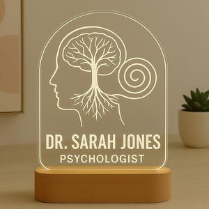 May include: A personalized desk lamp with a clear acrylic panel featuring a brain-tree design and the text "DR. SARAH JONES PSYCHOLOGIST." The lamp has a wooden base and emits a warm glow, suitable for a therapist's office.