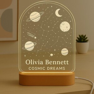May include: A personalized night light with a cosmic design. The acrylic panel features planets, stars, and constellations. The base is made of wood. The text on the light reads "Olivia Bennett" and "Cosmic Dreams".
