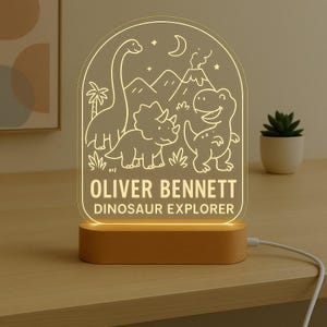 May include: An illuminated LED night light with a dinosaur design. The light has a wooden base and a clear acrylic panel with etched illustrations of dinosaurs, mountains, and the text "OLIVER BENNETT DINOSAUR EXPLORER."