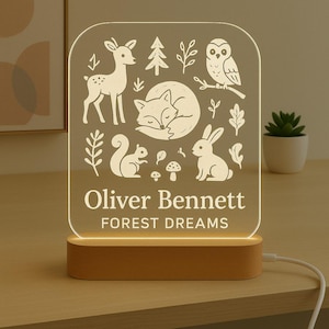 May include: A personalized night light with a wooden base and a clear acrylic panel. The panel features engraved forest animals, including a deer, fox, owl, squirrel, and rabbit. The text "Oliver Bennett FOREST DREAMS" is also engraved.