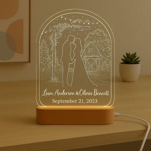 May include: A personalised LED night light with a wooden base. The clear acrylic panel features an etched design of a couple kissing in front of a barn. The names "Liam Anderson & Olivia Bennett" and the date "September 21, 2023" are displayed.