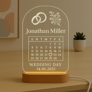 May include: A personalized wedding day LED lamp with a calendar design. The lamp features the names "Jonathan Miller" and the wedding date "14.06.2023". The design includes wedding rings and floral accents. The base is wooden.