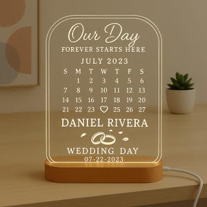 May include: A personalized wedding day LED night light. The clear acrylic panel displays the text "Our Day," a calendar for July 2023, names, and the wedding date. It sits on a wooden base and is lit with a warm glow.
