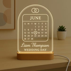 May include: A lighted acrylic wedding calendar with a wooden base. The calendar displays the month of June, with the 15th highlighted. The names "Liam Thompson" and "WEDDING DAY" are also displayed.