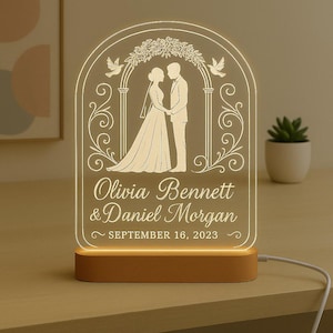 May include: A personalised wedding night light with a wooden base. The clear acrylic panel features an engraved image of a bride and groom under an arch, with the names "Olivia Bennett & Daniel Morgan" and the date "September 16, 2023".