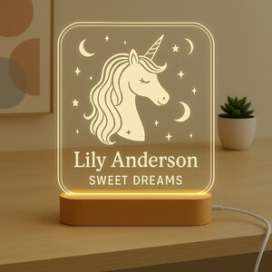 May include: A personalised night light with a unicorn design, stars, and crescent moons. The text "Lily Anderson SWEET DREAMS" is displayed on the illuminated panel, set on a wooden base. A white cord extends from the base.