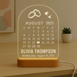 May include: A personalized wedding calendar night light with the text "AUGUST 2021" and "OLIVIA THOMPSON". The clear acrylic panel features a calendar, wedding rings, and birds, set on a wooden base.