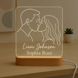 May include: A personalized LED night light featuring a line drawing of a couple kissing. The names "Liam Johnson" and "Sophia Rose" are inscribed below the image. The light sits on a rectangular wooden base.