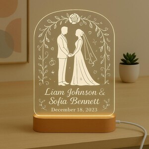 May include: A wedding-themed illuminated sign with a couple holding hands, framed by floral vines. The names "Liam Johnson & Sofia Bennett" and the date "December 18, 2023" are displayed. The base is light brown and the sign is lit.