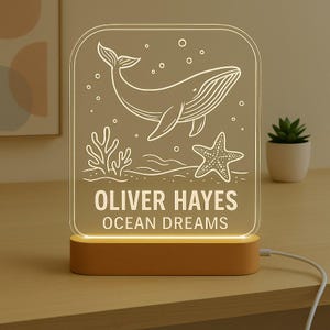 May include: A rectangular LED night light featuring a whale, starfish, and coral design. The light has the text "OLIVER HAYES OCEAN DREAMS" in white. The base is light brown, and the light is plugged in with a white cable.