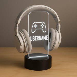 May include: A gaming-themed night light with a controller icon and the word "USERNAME" in white. The light is framed by grey headphones and sits on a black base. The light is designed for gamers.