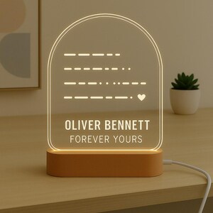 May include: A personalized night light with a warm glow. The clear acrylic panel features a Morse code message, a heart symbol, and the text "OLIVER BENNETT FOREVER YOURS." It sits on a wooden base and is plugged in.