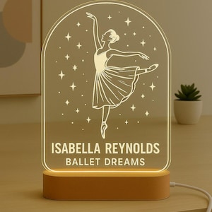 May include: A ballet-themed night light with a ballerina illustration and the text "ISABELLA REYNOLDS BALLET DREAMS" on a clear acrylic panel. The light sits on a wooden base and emits a warm glow, ideal for a dancer's bedroom. The measurements are in cm.