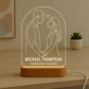 May include: An illuminated night light with a wooden base and a clear acrylic panel. The panel features an etched design of a couple holding hands, with the text "MICHAEL THOMPSON FOREVER YOURS." The light is connected with a white cable.