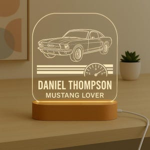 May include: A personalised Mustang-themed night light. The clear acrylic panel features a car illustration, speedometer graphic, and the text "DANIEL THOMPSON MUSTANG LOVER." It sits on a wooden base and is illuminated.