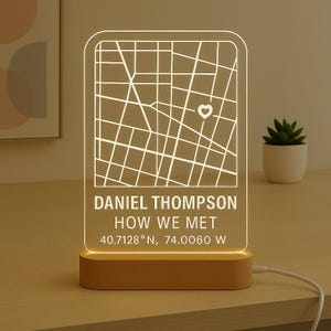 May include: A rectangular LED lamp with a map design, a heart icon, and the text "DANIEL THOMPSON HOW WE MET 40.7128° N, 74.0060 W". The lamp has a wooden base and a white cord.