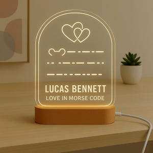 May include: A personalized LED night light with a wooden base. The clear acrylic panel features two interlocking hearts, Morse code, and the text "LUCAS BENNETT LOVE IN MORSE CODE". The light is plugged in with a white cord.