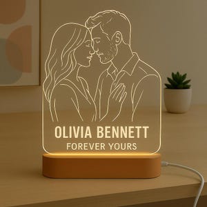 May include: An illuminated LED night light with a couple's outline, displaying the names "OLIVIA BENNETT" and the phrase "FOREVER YOURS" in white. The light is set on a wooden base and is plugged in.