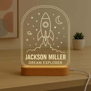 May include: An illuminated LED night light with a rocket ship design, stars, a crescent moon, and the text "JACKSON MILLER DREAM EXPLORER." It has a wooden base and a white power cable.