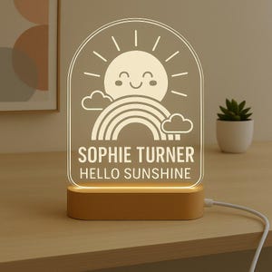 May include: A personalised night light featuring a smiling sun, rainbow, and clouds design. The text "SOPHIE TURNER HELLO SUNSHINE" is displayed below the graphic. The light sits on a wooden base and is plugged in.