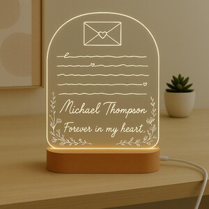 May include: A lighted acrylic memorial plaque with a wooden base. The plaque features an engraved envelope design, lines of text, and the words "Michael Thompson Forever in my heart." The plaque is illuminated with a warm glow.