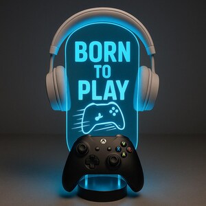 May include: A gaming-themed LED lamp with the text "BORN TO PLAY" in white. The lamp has a blue glow and features a controller graphic. A black game controller sits below the lamp, and white headphones are positioned above.