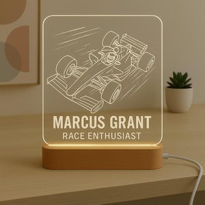 May include: A personalized LED night light featuring a race car design. The light has a wooden base and a clear acrylic panel with the name "MARCUS GRANT" and the text "RACE ENTHUSIAST" etched on it. The light is plugged into a power source.