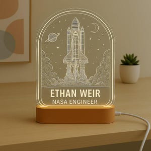 May include: A personalized night light featuring a space shuttle design with the name "ETHAN WEIR" and the title "NASA ENGINEER." The light has a warm glow and sits on a rectangular base. The design includes a planet, stars, and a crescent moon.