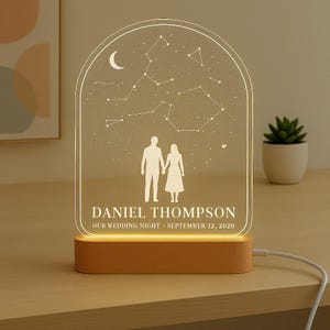 May include: A personalised night light featuring a constellation design, a crescent moon, and a couple holding hands. The text reads "DANIEL THOMPSON, OUR WEDDING NIGHT, SEPTEMBER 12, 2020." The light has a wooden base and a clear acrylic top.