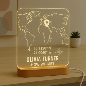 May include: A personalised LED night light with a world map design. The light features a heart-shaped pin, coordinates, the names "Olivia Turner," and the phrase "How We Met." The base is made of wood.