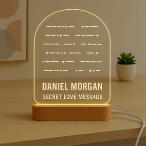 May include: A personalised night light with the name "DANIEL MORGAN" and the text "SECRET LOVE MESSAGE" illuminated on a clear acrylic panel. The panel features a Morse code design and sits on a wooden base.