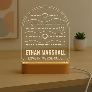 May include: A personalised LED night light with a wooden base. The clear acrylic panel displays the name "ETHAN MARSHALL" and the phrase "LOVE IN MORSE CODE" in white. The design includes hearts and Morse code symbols.