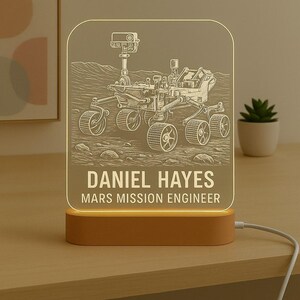 May include: An illuminated night light with a Mars rover design. The light is engraved with the name "DANIEL HAYES" and the title "MARS MISSION ENGINEER". The base is a warm brown colour, and the light emits a soft glow, perfect for a desk or bedside table.