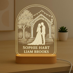 May include: A personalised wedding night light showing a couple under an archway, with the names "SOPHIE HART" and "LIAM BROOKS" engraved. The light sits on a wooden base and emits a warm glow. The design includes a tree and fairy lights.