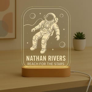 May include: A personalized LED night light featuring an astronaut illustration. The light has a wooden base and a clear acrylic panel with the name "NATHAN RIVERS" and the text "REACH FOR THE STARS". The astronaut is floating in space.