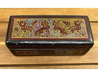 May include: A rectangular wooden box with a black exterior and a burgundy top. The top features a gold and burgundy design of stylized creatures. The box is likely used for storing small items.