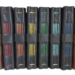 May include: A row of eight black leather-bound journals with colorful stitching. The stitching is in different colors, including red, orange, yellow, green, blue, and turquoise.