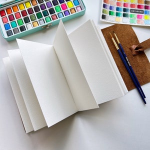May include: An open watercolor sketchbook with blank pages, a brown leather cover, and two paintbrushes. The sketchbook is lying on a white surface.