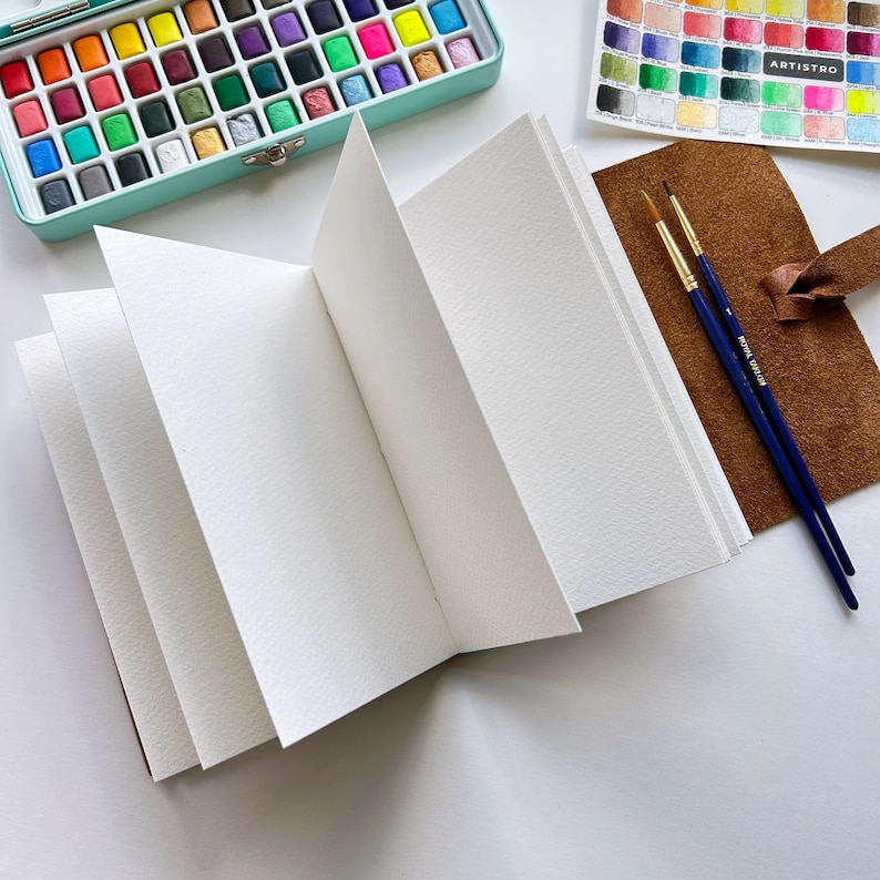 May include: An open watercolor sketchbook with blank pages, a brown leather cover, and two paintbrushes. The sketchbook is lying on a white surface.