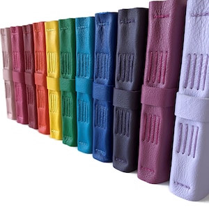 May include: A row of ten leather-bound journals in various colors, including purple, pink, lavender, red, orange, yellow, green, blue, and navy. Each journal has a stitched spine and a leather strap.