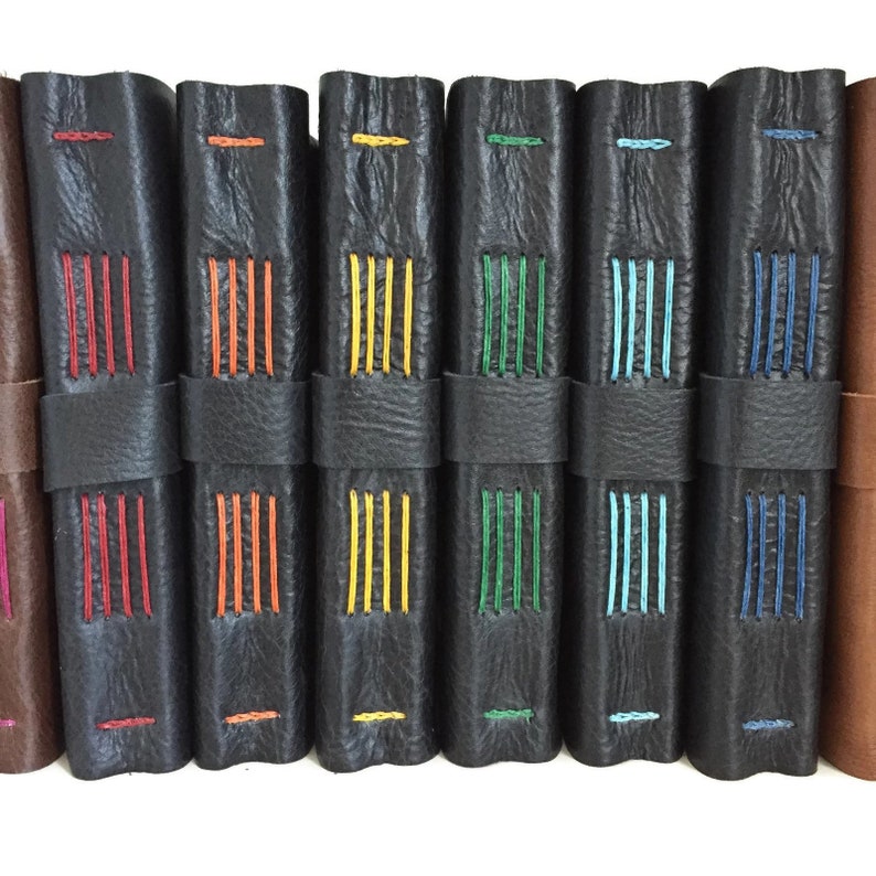 May include: A row of eight black leather-bound journals with colorful stitching. The stitching is in different colors, including red, orange, yellow, green, blue, and turquoise.