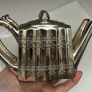 May include: Silver teapot with a geometric design. The teapot features a curved handle, a spout, and a lid with a decorative finial. The body of the teapot is vertically ribbed with etched detailing. The teapot is held by a hand.